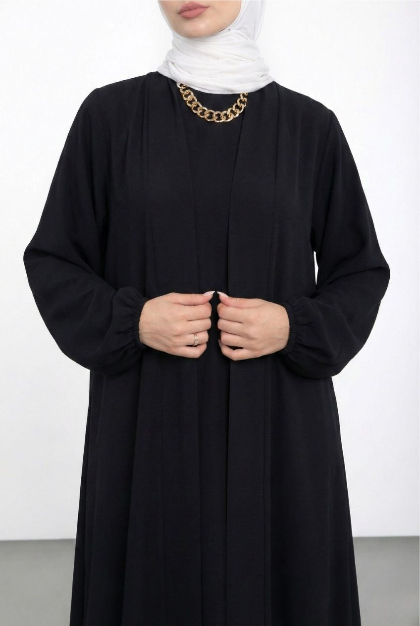 Person wearing a black abaya with a white hijab and gold necklace on a white background