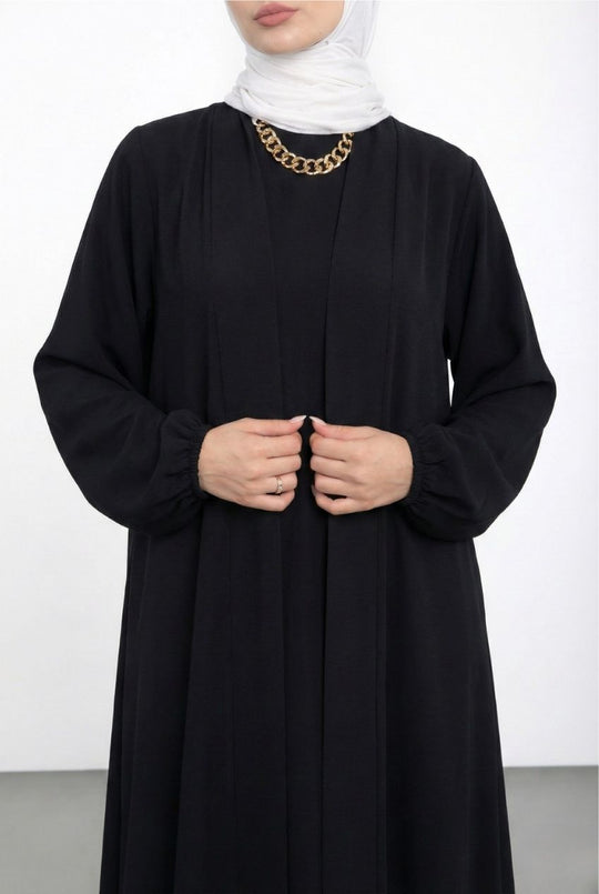 Person wearing a black abaya with a white hijab and gold necklace on a white background