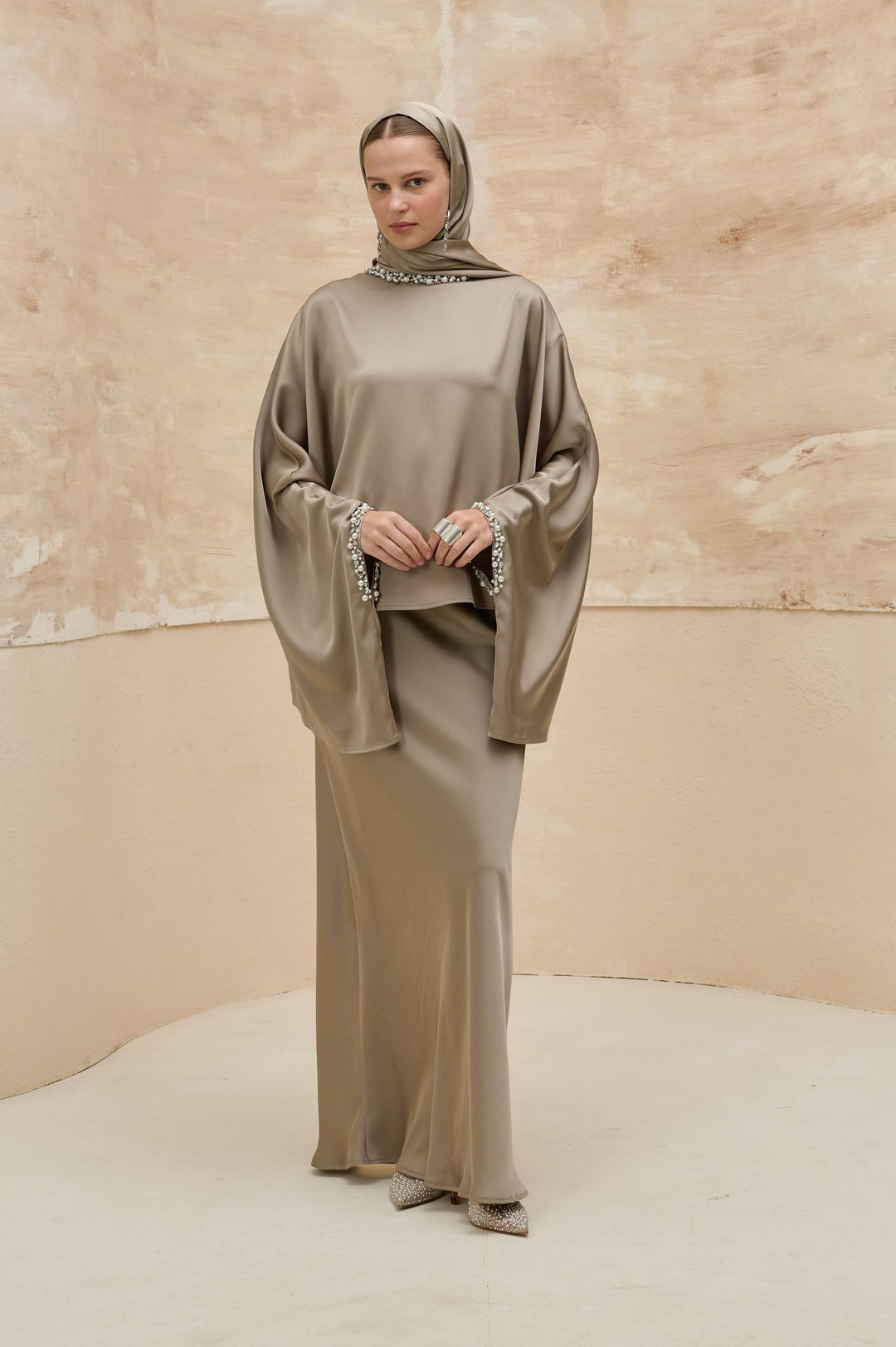 Woman wearing a long, light brown dress with a matching headscarf against a beige background