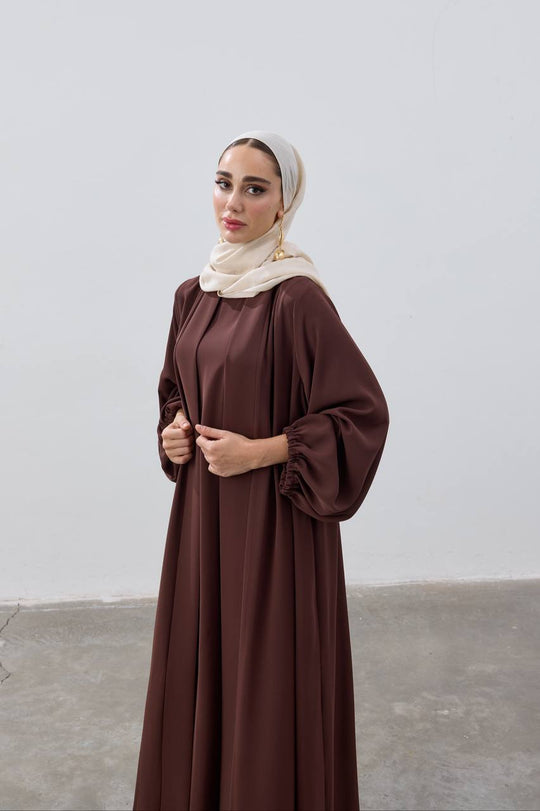Woman wearing a brown dress and beige hijab against a plain background