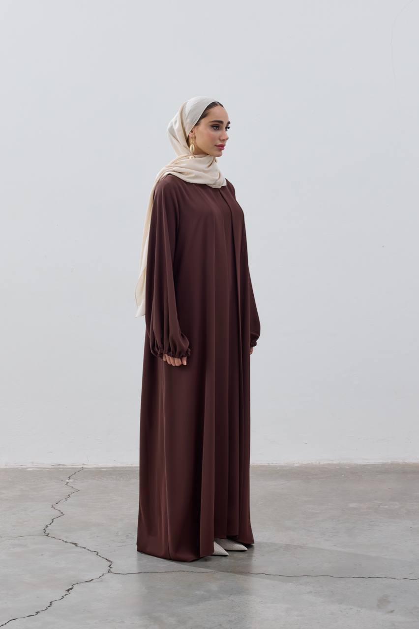 Woman wearing a brown dress and beige hijab standing against a white wall.