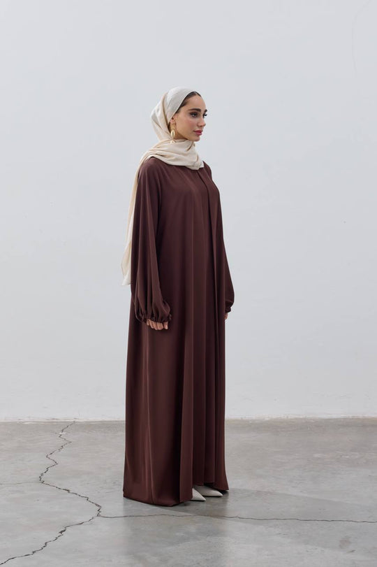 Woman wearing a brown dress and beige hijab standing against a white wall.