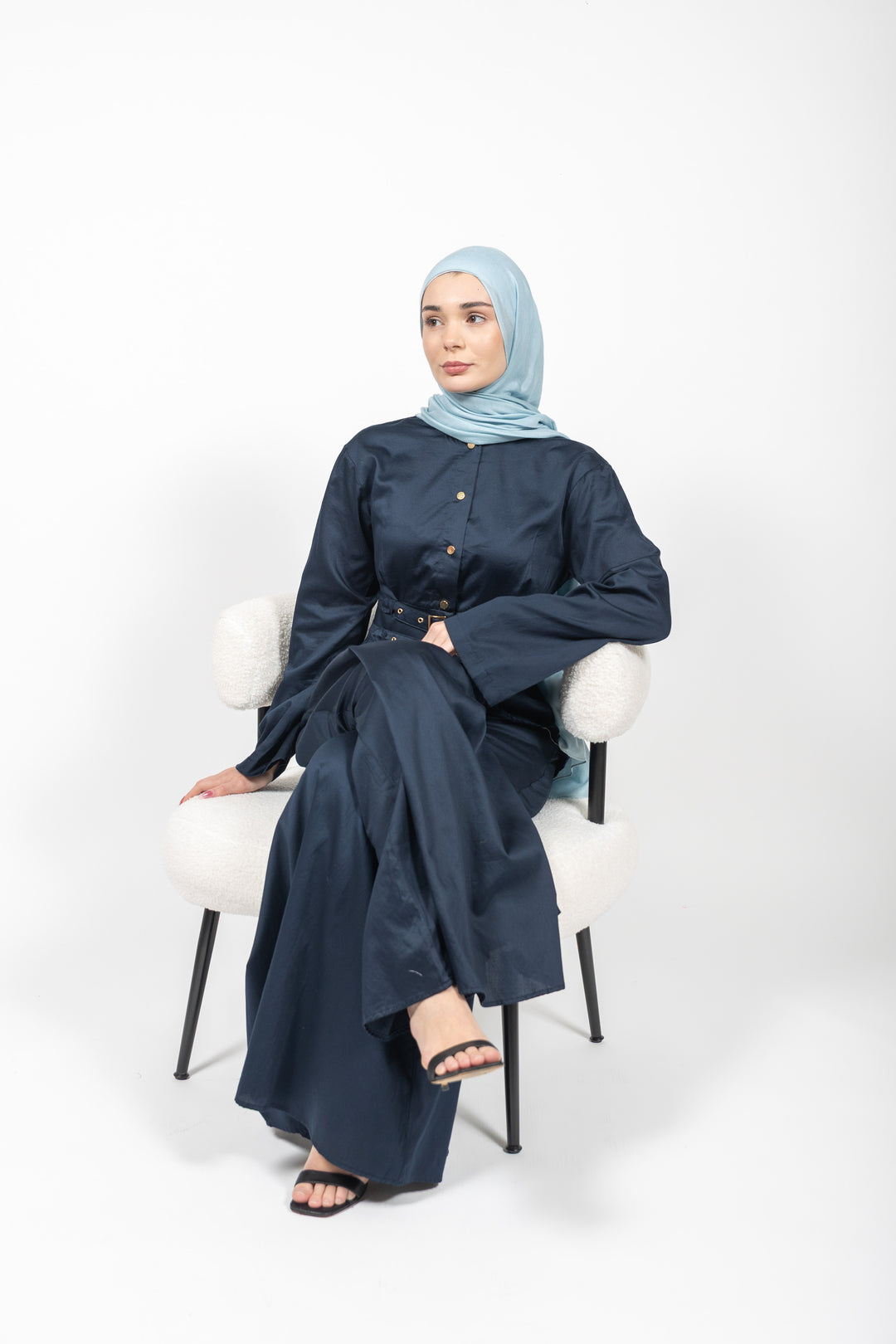 Woman wearing a navy blue outfit with a light blue hijab sitting on a white chair against a white background