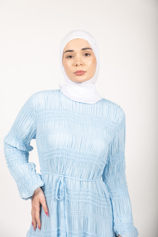 Woman wearing a light blue dress with long sleeves and a white hijab on a plain background