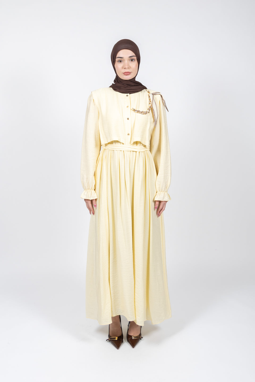 Woman wearing a light yellow dress with long sleeves and a brown hijab on a white background