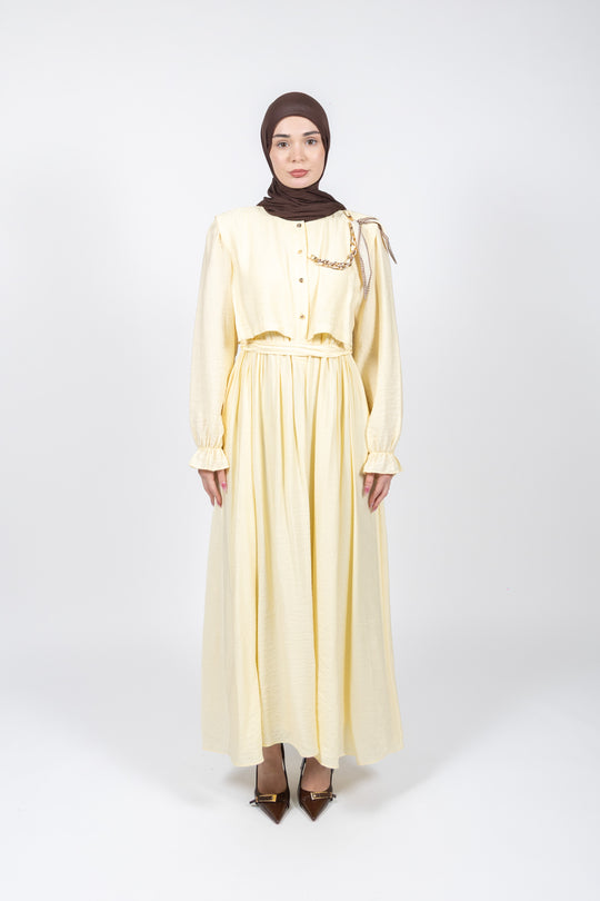 Woman wearing a light yellow dress with long sleeves and a brown hijab on a white background